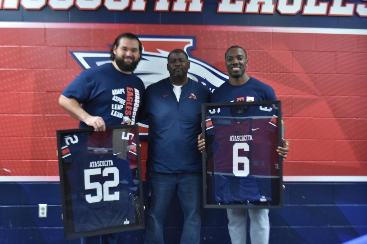 Alumni Samuel Cosmi and Patrick Taylor stand with principal Falker as they hold their retired jerseys.